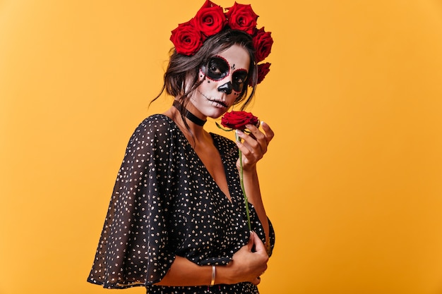 Ritratto di ragazza calma con capelli scuri in corona di fiori rossi. Donna con maschera scheletro godendo profumo di fiori.