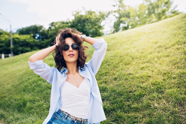 Ritratto di ragazza carina bruna con i capelli corti in occhiali da sole in posa per la macchina fotografica nel parco su sfondo prato. Indossa una maglietta bianca, una maglietta blu.