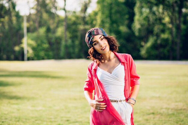 Ritratto di ragazza carina bruna in bandana in piedi nel parco estivo. Indossa abiti bianchi, una lunga camicia rosa. Sta guardando alla telecamera.