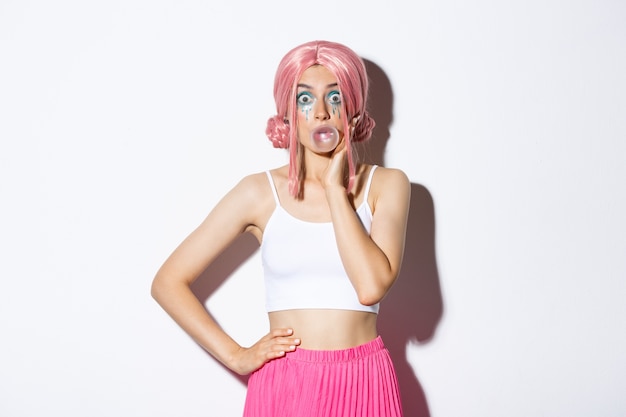 Ritratto di ragazza carina sorpresa in parrucca rosa, che soffia gomma da masticare e guardando stupito alla telecamera, in piedi.