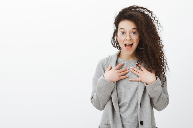 Ritratto di ragazza entusiasta sorpresa con capelli ricci in occhiali trasparenti alla moda e cappotto grigio, con i palmi delle mani sul petto, ansimante e sorridente ampiamente con la bocca aperta