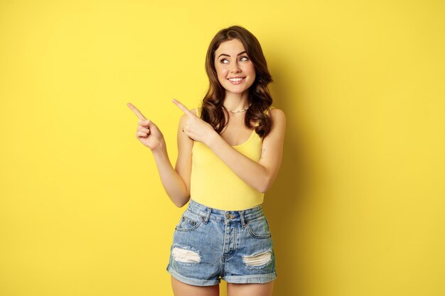 Ritratto di ragazza estiva alla moda, donna bruna che mostra il modo, puntando il dito a sinistra, pubblicità, in piedi su sfondo giallo. Copia spazio