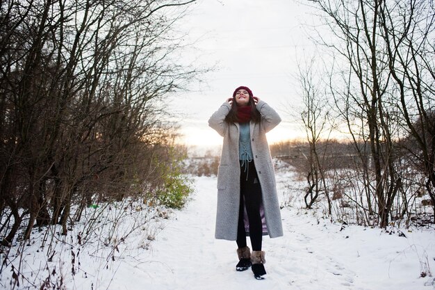 Ritratto di ragazza gentile con cappello rosso cappotto grigio e sciarpa vicino ai rami di un albero innevato