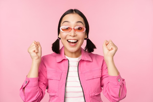Ritratto di ragazza giapponese eccitata in occhiali da sole che celebra il raggiungimento dell'obiettivo ansimante stupito e sorridente in piedi su sfondo rosa