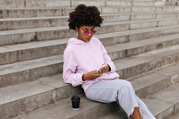 Ritratto di ragazza riccia allegra in pantaloni di jeans, occhiali da sole rosa e felpa con cappuccio viola che tiene il telefono e si siede sulle scale all'aperto