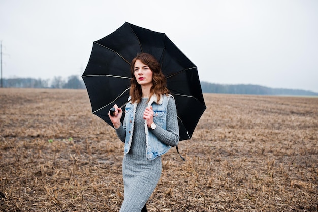 Ritratto di ragazza riccia bruna in giacca di jeans con ombrello nero al campo
