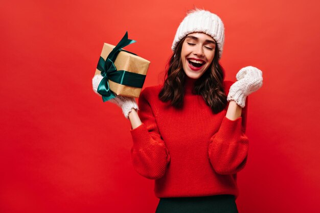 Ritratto di ragazza riccia eccitata con cappello bianco e guanti lavorati a maglia si rallegra e tiene una confezione regalo su sfondo rosso isolato