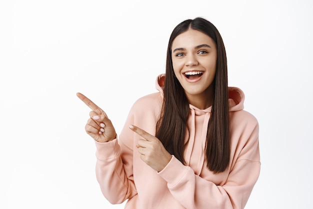 Ritratto di ragazza sorridente allegra con i capelli lunghi che puntano le dita nell'angolo in alto a sinistra gioiosa che mostra banner logo in piedi su sfondo bianco