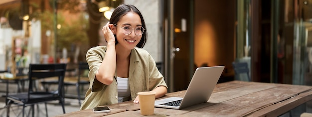 Ritratto di ragazza sorridente con gli occhiali seduta con il computer portatile in un bar all'aperto, bere caffè e lavorare