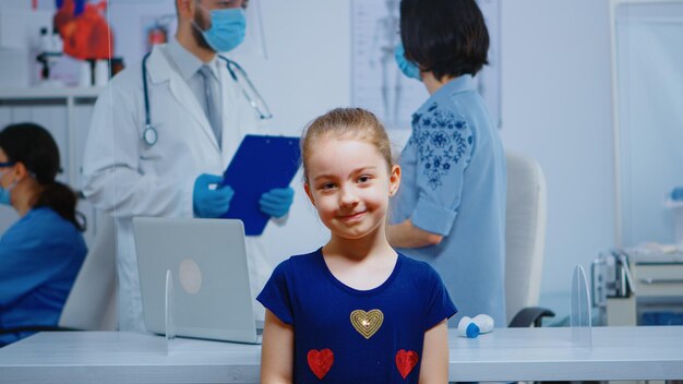 Ritratto di ragazza sorridente in studio medico mentre la madre parla con il medico in background. Specialista in medicina con maschera di protezione che fornisce servizi di assistenza sanitaria, consulenza in clinica ospedaliera