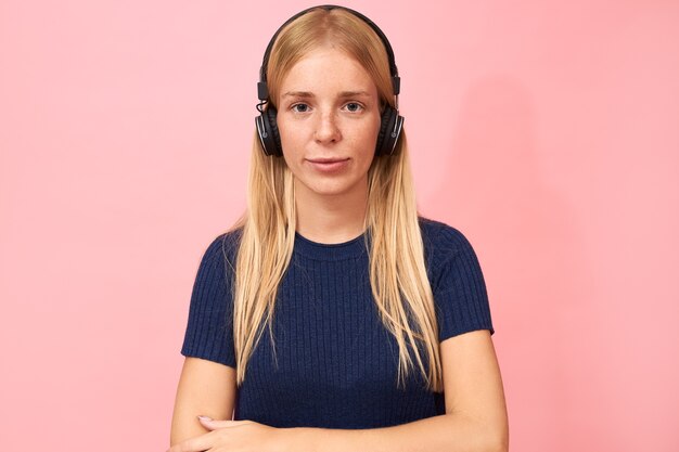 Ritratto di ragazza studentessa alla moda con anello al naso in posa sul rosa in cuffie wireless, ascoltando lezioni online, audiolibri o podcast