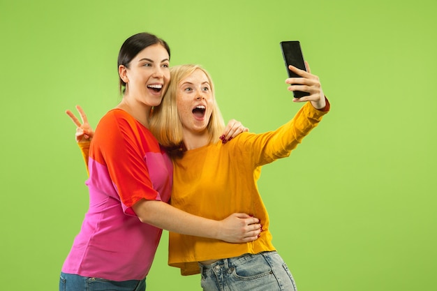 Ritratto di ragazze abbastanza affascinanti in abiti casual isolati su sfondo verde studio. Fidanzate o lesbiche che fanno selfie. Concetto di LGBT, uguaglianza, emozioni umane, amore, relazione.