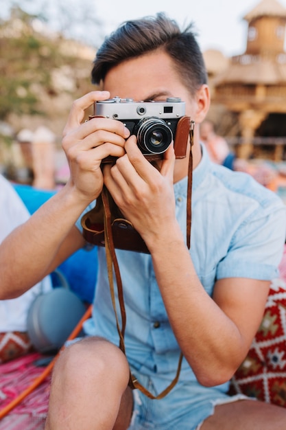 Ritratto di ragazzo con capelli neri corti, tenendo la fotocamera retrò e fare foto, mentre è seduto in un caffè all'aperto