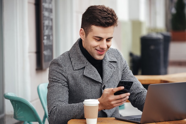 Ritratto di ragazzo di successo che riposa in street cafe, lavorando con il notebook e digitando il messaggio di testo sul suo telefono cellulare