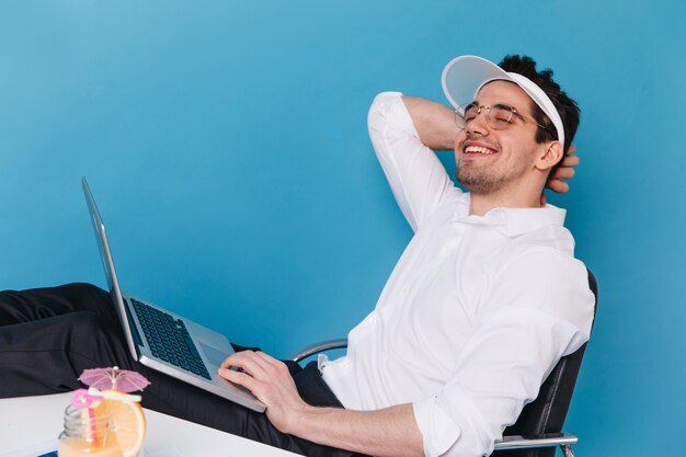 Ritratto di ragazzo gioioso in bicchieri, cappello bianco e camicia sorridente e tenendo il computer portatile.