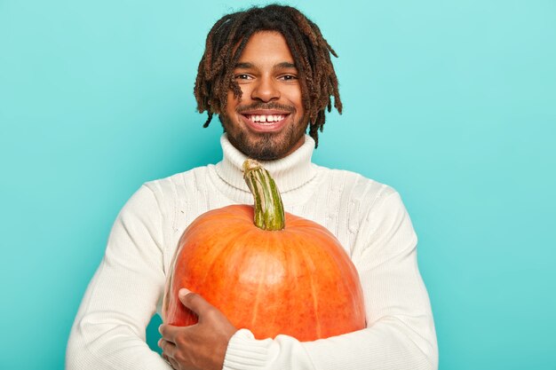 Ritratto di ragazzo sorridente felice ha umore felice, trasporta grande zucca arancione, indossa un maglione bianco, isolato su sfondo blu.