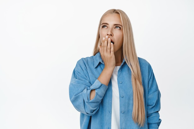 Ritratto di sbadigliare ragazza bionda che copre la bocca con la mano e alzando lo sguardo annoiato in piedi stanco su sfondo bianco