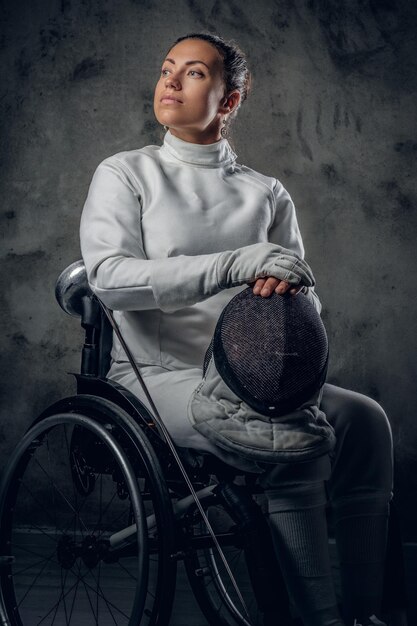 Ritratto di schermidore femminile in sedia a rotelle con maschera di sicurezza e pinza su sfondo grigio.