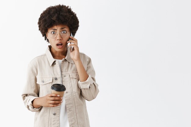 Ritratto di scosso donna afroamericana confusa e stordita in bicchieri e camicia elegante, bere caffè e parlare al telefono