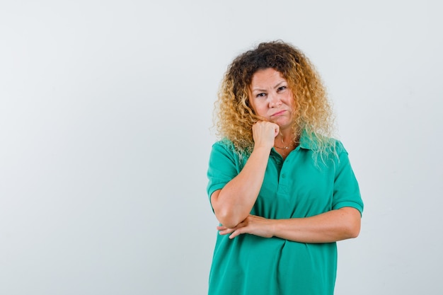 Ritratto di signora abbastanza bionda che sostiene il mento a portata di mano in maglietta polo verde e guardando vista frontale delusa