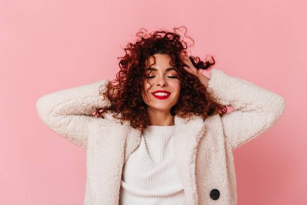 Ritratto di signora affascinante con rossetto rosso e sorriso bianco come la neve vestita con un cappotto ecologico luminoso e toccando i suoi capelli ricci sullo spazio rosa.