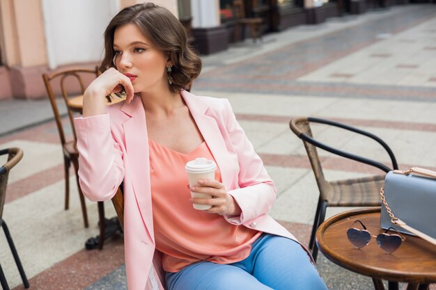 Ritratto di signora pensante alla moda seduta a tavola a bere caffè in giacca rosa tendenza stile estivo, borsa blu, accessori, street style, moda donna