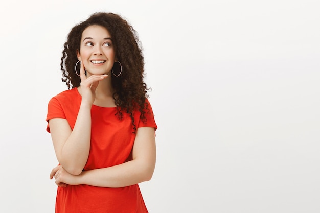 Ritratto di sognante bella donna elegante con i capelli ricci in abito rosso, sorridente con gioia mentre guarda da parte, tenendo la mano sul mento