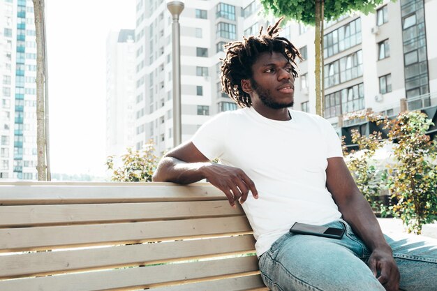 Ritratto di sorridente bel modello hipsterUnshaven africano uomo vestito con maglietta bianca estiva e jeans Moda maschile con acconciatura di dreadlocks in posa sullo sfondo della strada