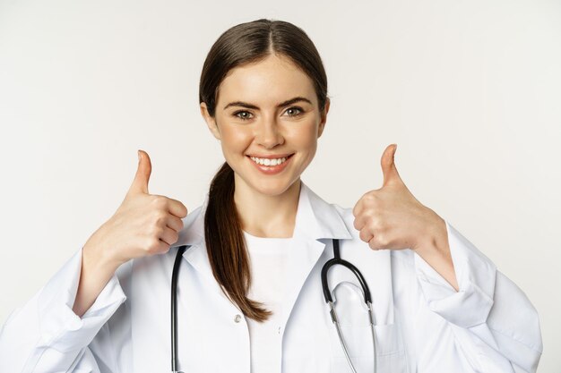 Ritratto di sorridente bella donna medico in camice bianco che mostra i pollici in su in piedi su sfondo bianco...