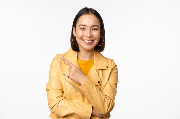 Ritratto di sorridente ragazza asiatica che punta il dito a sinistra mostrando il logo o copia la pubblicità dello spazio sul lato vuoto in piedi su sfondo bianco
