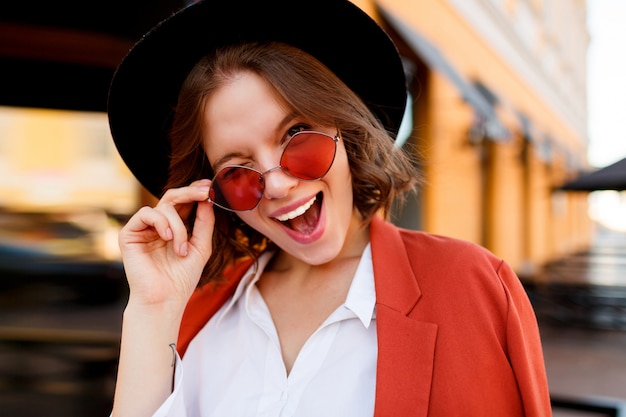 Ritratto di sorridente ragazza europea in simpatici occhiali da sole arancione, giacca e cappello nero. Moda autunno. Caffè all'aperto.