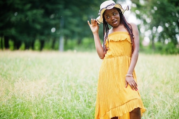 Ritratto di splendida donna afroamericana anni '20 in abito giallo e cappello estivo in posa all'erba verde nel parco