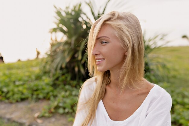 Ritratto di splendida donna europea anni '20 con i capelli lunghi in abito bianco estivo che ride e distoglie lo sguardo mentre si cammina sulla sabbia in riva al mare