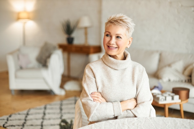 Ritratto di splendida femmina europea matura con acconciatura corta rilassante a casa seduto al tavolo in soggiorno incrociando le braccia sul petto cercando di riscaldarsi in maglione dolcevita di cashmere accogliente, sorridente