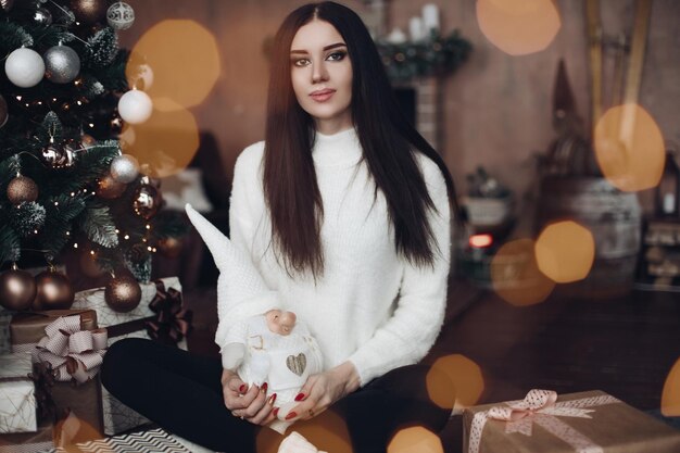 Ritratto di splendida giovane donna attraente con lunghi capelli scuri e trucco da sera luminoso in maglione bianco seduto sul pavimento con regali di Natale Sta tenendo in mano un nano giocattolo bianco Guardando la fotocamera