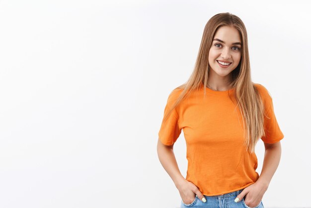 Ritratto di splendida studentessa bionda ragazza in maglietta arancione sorridente fotocamera entusiasta con bella espressione felice in piedi su sfondo bianco posa rilassata con le mani in tasca