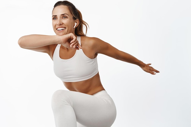 Ritratto di sportiva che allunga il corpo che si esercita facendo fitness gamba alzare e sorridente in esecuzione in piedi su sfondo bianco