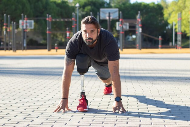 Ritratto di sportivo serio con disabilità sul campo sportivo. Uomo bello che si allena allo stadio tenendo le mani sul marciapiede che si prepara a correre. Stile di vita sano delle persone con concetto di disabilità