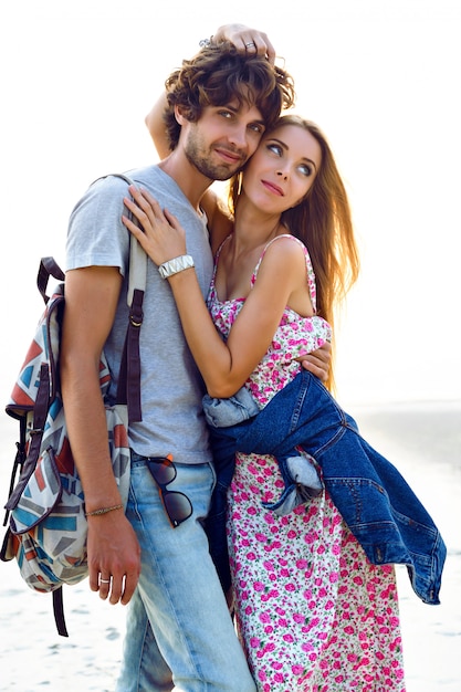 Ritratto di stile di vita all'aperto di incredibili coppie abbastanza giovani innamorate in posa sulla spiaggia. Elegante uomo e donna abbraccia e trascorrono bei momenti insieme. vestito floreale zaino e denim.