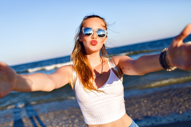 Ritratto di stile di vita all'aperto di una ragazza felice divertente che viaggia da sola verso l'oceano, facendo selfie in spiaggia, emozioni positive felici, occhiali da sole a specchio, top corto bianco e zaino, gioia, movimento.