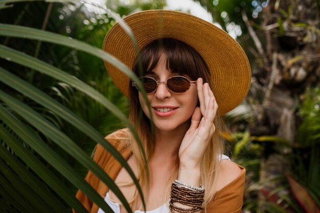 Ritratto di stile di vita della donna castana in cappello di paglia che posa sulle foglie di palma tropicale