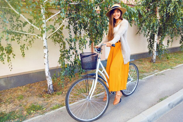 Ritratto di stile di vita di moda all'aperto di bella giovane ragazza bruna in sella alla sua bicicletta retrò sulla strada con alberi di betulla. Indossare abiti eleganti e alla moda, cappello e cardigan caldo. Umore autunnale.