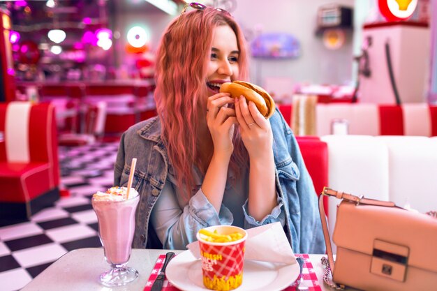 Ritratto di stile di vita positivo di donna graziosa uscita felice con i capelli rosa a cena al caffè americano vintage, mangiare hot dog, patatine fritte e mil shake, cheat meal di cibo spazzatura, colori pastello.