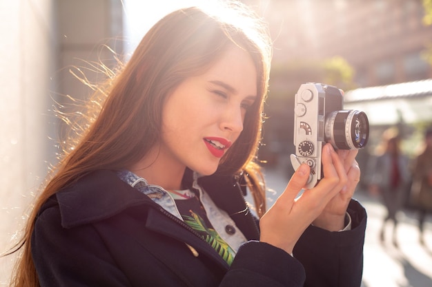 Ritratto di stile di vita sorridente all'aperto di una giovane donna piuttosto giovane, divertendosi in città con la fotocamera, foto di viaggio del fotografo. Fare foto in stile hipster. Bagliore del sole