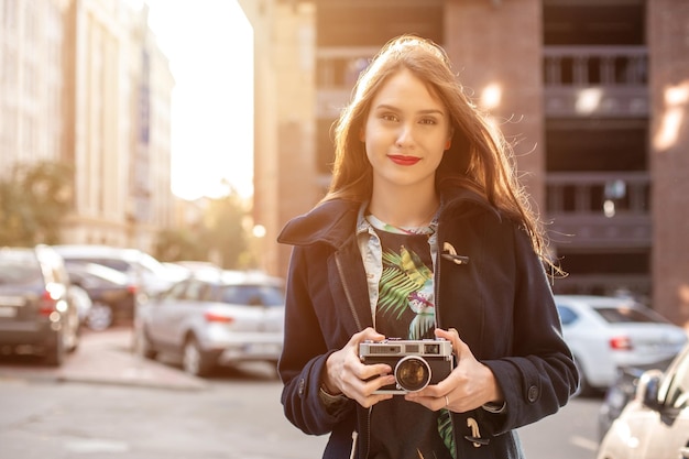 Ritratto di stile di vita sorridente all'aperto di una giovane donna piuttosto giovane, divertendosi in città con la fotocamera, foto di viaggio del fotografo. Fare foto in stile hipster. Bagliore del sole