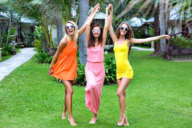 Ritratto di stile di vita tropicale estivo di tre ragazze felici dei migliori amici che si divertono all'aperto, indossando abiti sexy colorati, vacanza in stile spiaggia, giardino esotico, occhiali da sole vestiti alla moda, relax, gioia