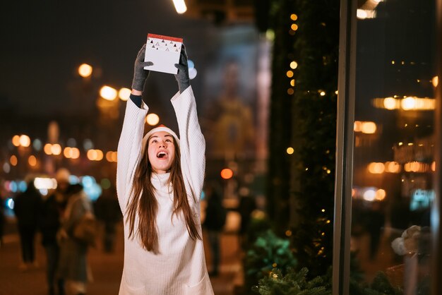 Ritratto di strada di notte di giovane bella donna che agisce entusiasta. Luci festive della ghirlanda.