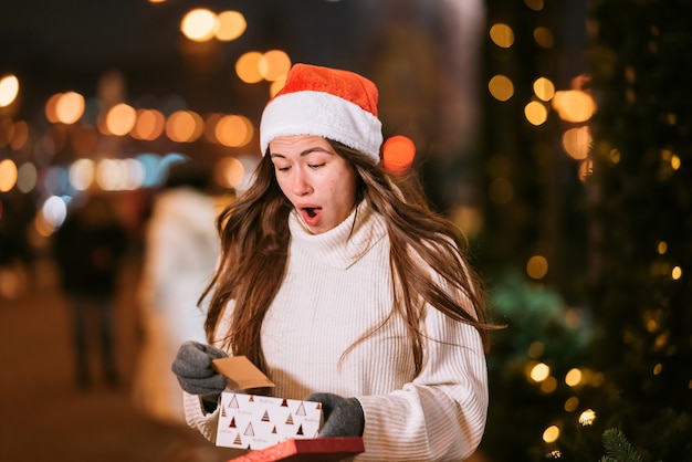 Ritratto di strada di notte di giovane bella donna che agisce entusiasta. Luci festive della ghirlanda.