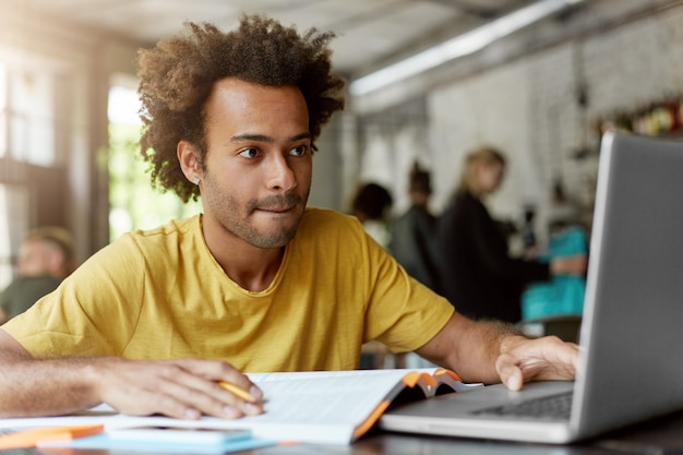 Ritratto di studente intelligente con pelle scura e capelli folti che indossa abiti casual mentre è seduto alla mensa che lavora al suo documento di corso alla ricerca di informazioni in Internet utilizzando il suo computer portatile