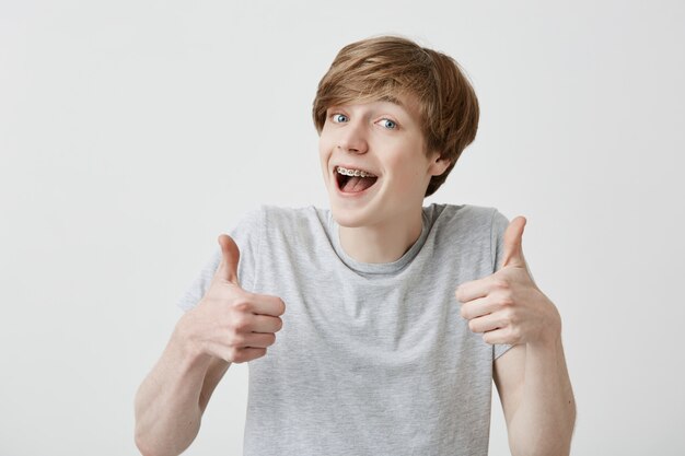 Ritratto di studente maschio biondo o cliente con ampio sorriso, guardando la telecamera con espressione felice, mostrando il pollice in su con entrambe le mani, raggiungendo obiettivi di studio. Linguaggio del corpo